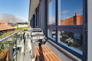 A balcony with a barbecue grill, a wooden bench, and a glass railing overlooking a residential area. - Cocomo Stylish Pet Friendly Elwood Apartment (Elwood)