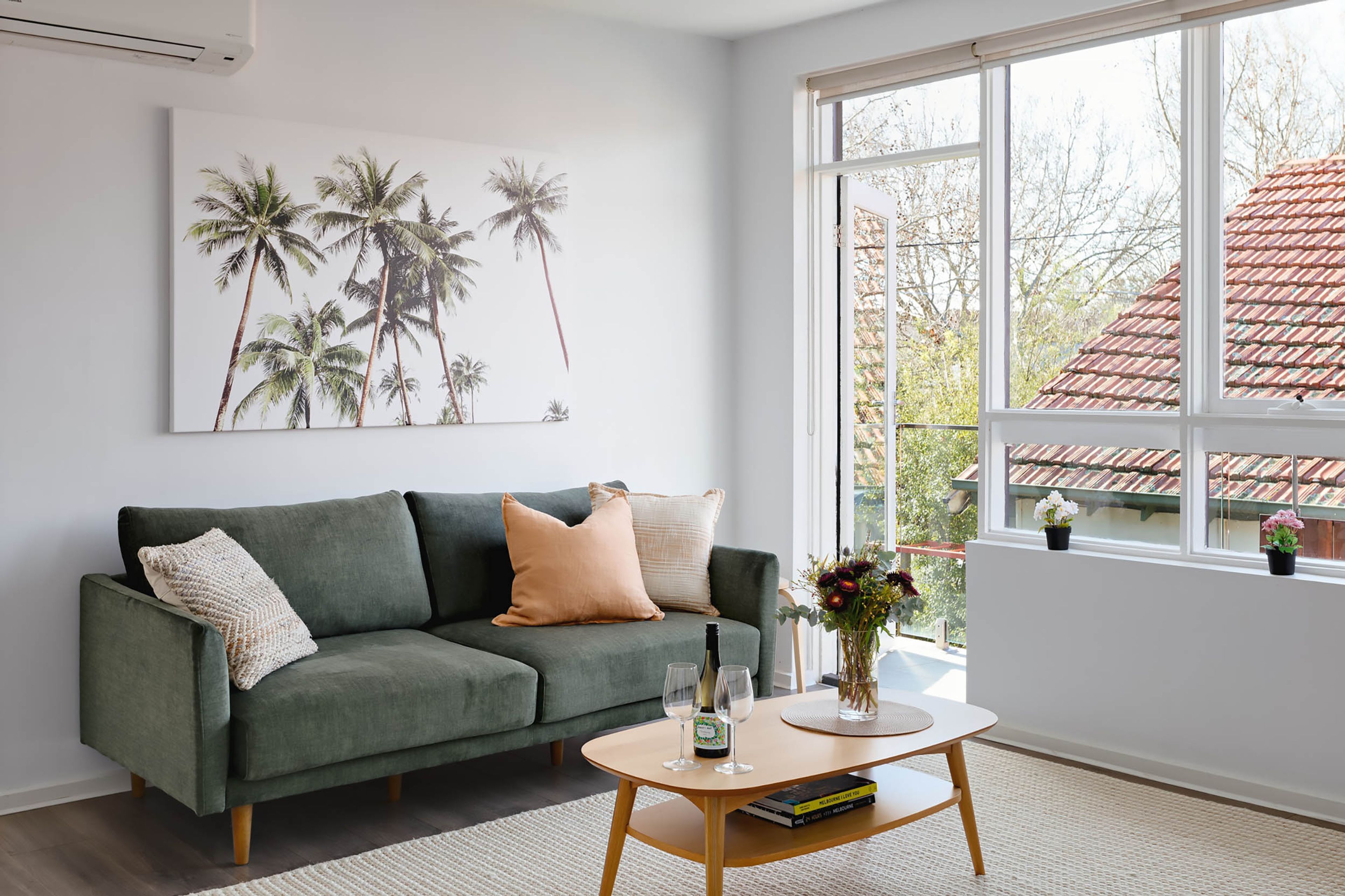 A modern living room with a green sofa, a coffee table, and a large window with a view of a red-tiled roof.