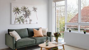 A modern living room with a green sofa, a coffee table, and a large window with a view of a red-tiled roof. - Cocomo Stylish Pet Friendly Elwood Apartment (Elwood)