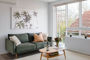 A modern living room with a green sofa, a coffee table, and a large window with a view of a red-tiled roof. - Cocomo Stylish Pet Friendly Elwood Apartment (Elwood)