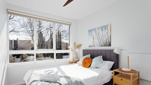 Une chambre avec une grande fenêtre, un lit avec une literie blanche, une tête de lit grise et une table de chevet avec une lampe.