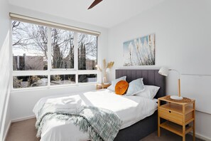 A bedroom with a large window, a bed with white bedding, a grey headboard, and a bedside table with a lamp. - Cocomo Stylish Pet Friendly Elwood Apartment (Elwood)