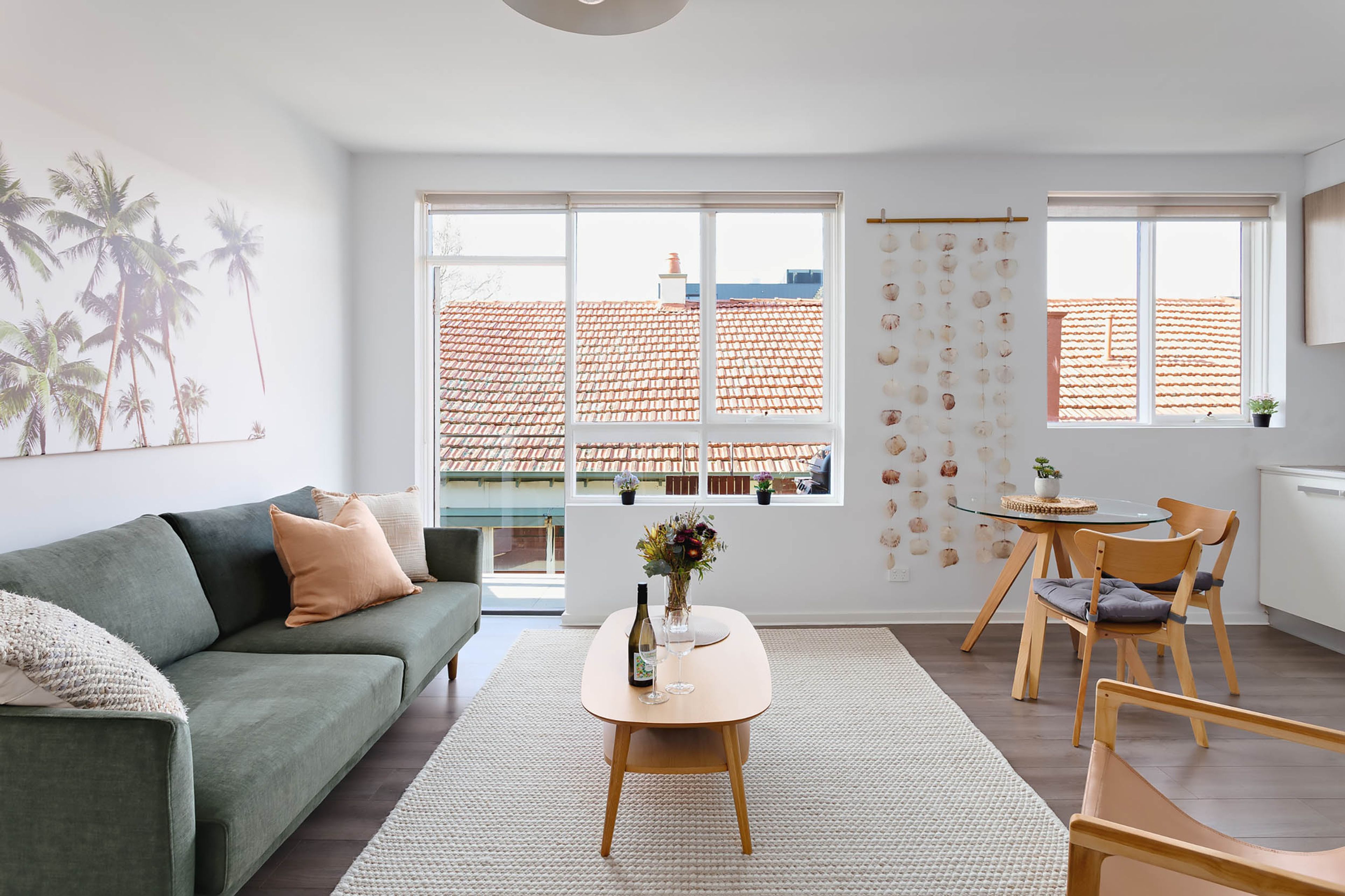Un salon moderne comprenant un canapé, une table basse avec un vase, un coin repas avec une table et des chaises, de grandes fenêtres offrant une vue sur un toit recouvert de tuiles rouges, et un mur orné de décorations suspendues.