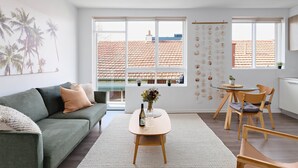 Un salon moderne comprenant un canapé, une table basse avec un vase, un coin repas avec une table et des chaises, de grandes fenêtres offrant une vue sur un toit recouvert de tuiles rouges, et un mur orné de décorations suspendues.