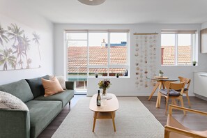 A modern living room with a sofa, a coffee table with a vase, a dining area with a table and chairs, large windows with a view of a red-tiled roof, and a wall with hanging decorations. - Cocomo Stylish Pet Friendly Elwood Apartment (Elwood)