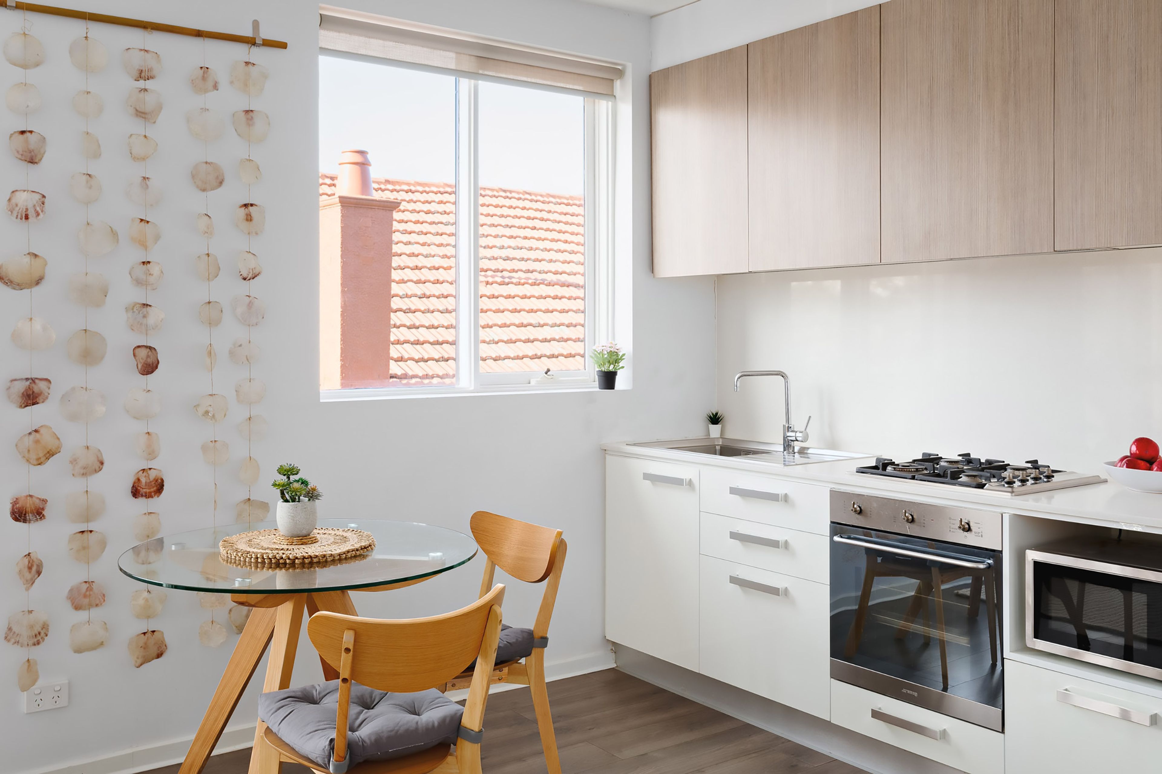 A modern kitchen with white cabinets, a gas stove, and a built-in oven. There is a round glass table with a cushion and a chair.