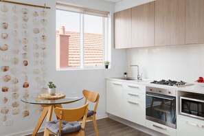 Une cuisine moderne avec des meubles blancs, une cuisinière à gaz et un four encastré. On y trouve une table ronde en verre, un coussin et une chaise.