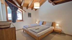 A bedroom with a wooden bed, two bedside tables with lamps, a window with curtains, and a wooden dresser. - Apartment Alpina (Zermatt)
