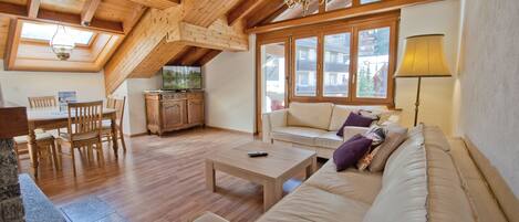 A spacious living area with a wooden ceiling, a sofa set, a dining table with chairs, and a television.