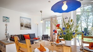 A modern living room with a wooden dining table, grey sofas, and a large window with a view of greenery. - Apartment for up to 4 people,central,quiet,south-facing terrace,sauna,WLAN,bicycle shed,parking lot (Prerow)