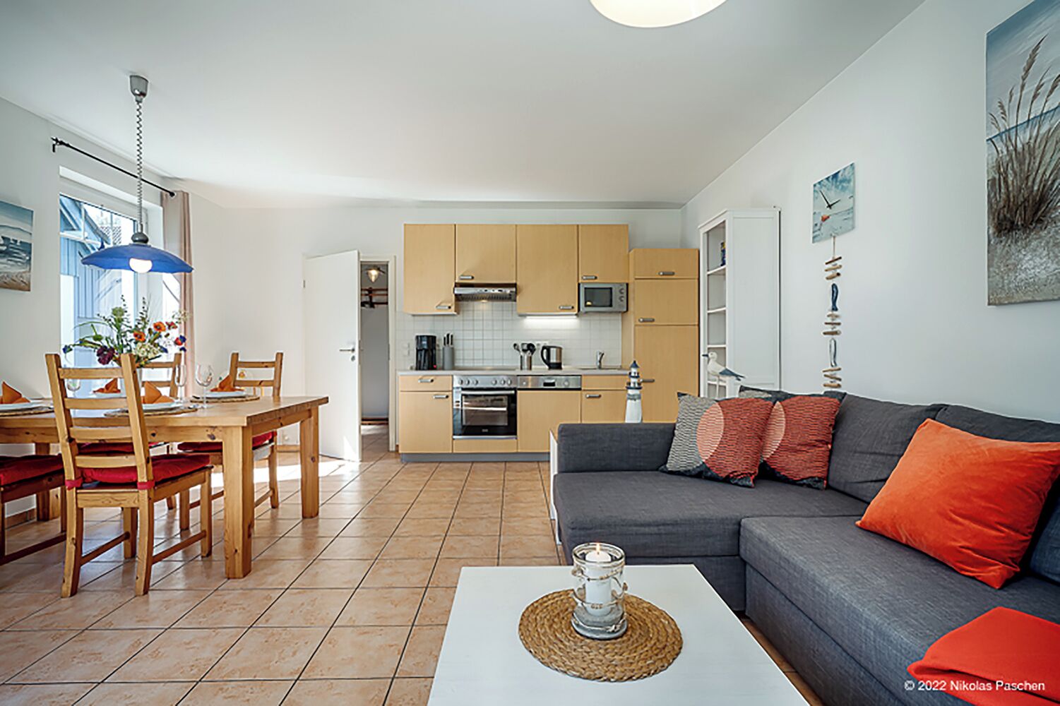 A modern living room with a grey sofa, red cushions, a dining table with chairs, and a kitchen in the background.
