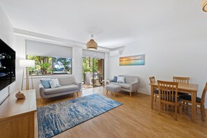 A spacious living room with a sofa, coffee table, and dining table. There is a large window providing a view of the outdoors. - Flynn's Beach Getaway (Port Macquarie)