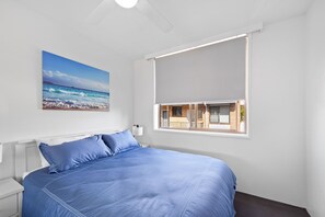 Un dormitorio con una cama grande, una ventana con vista a un edificio y un cuadro de una playa.