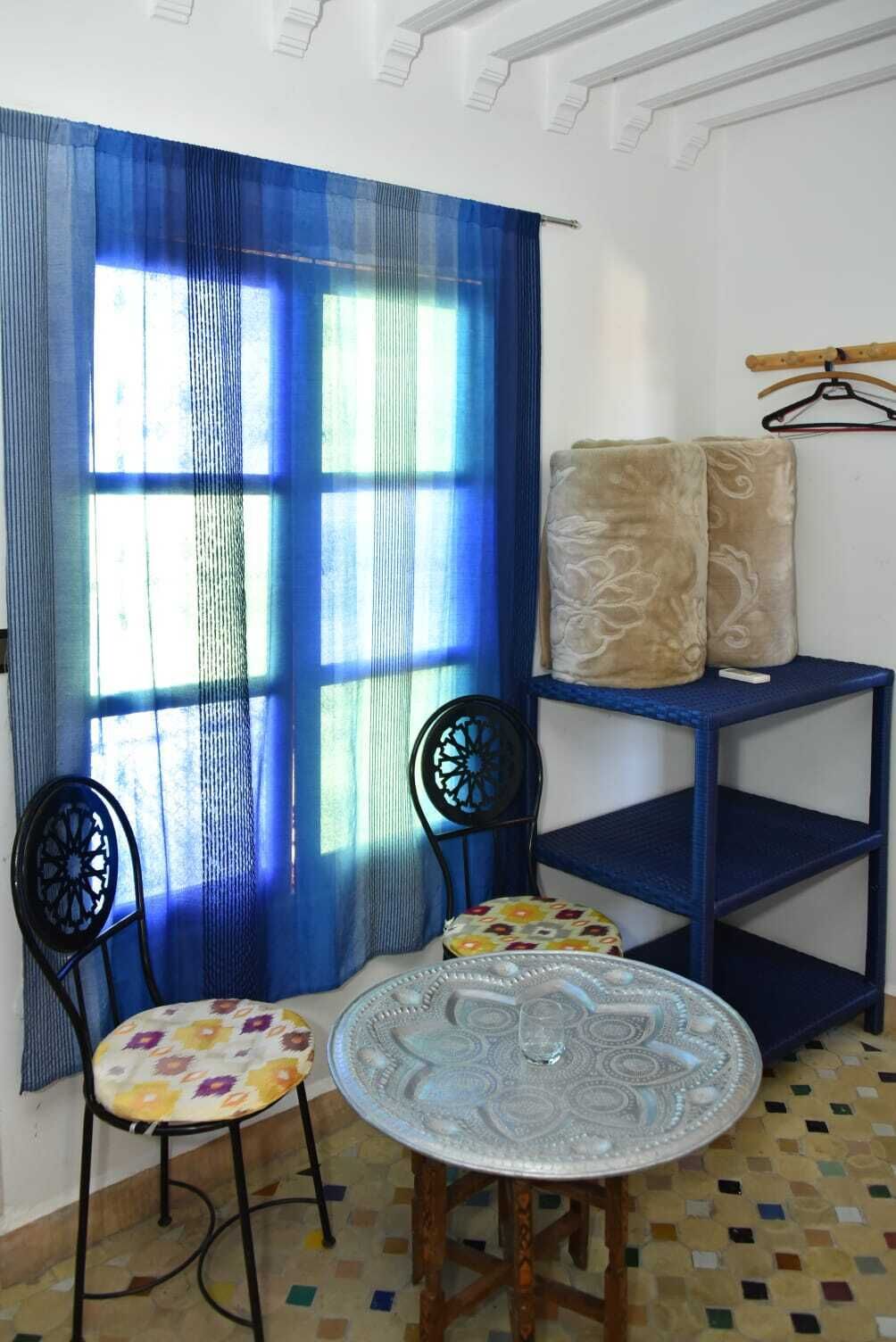 Una habitación con una cortina azul, una mesa redonda con superficie de vidrio, dos sillas, un pequeño estante con cojines y un perchero.