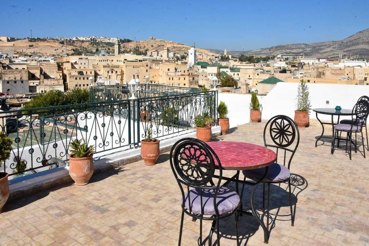 A terrace with outdoor seating, potted plants, and a view of a cityscape with buildings and hills.