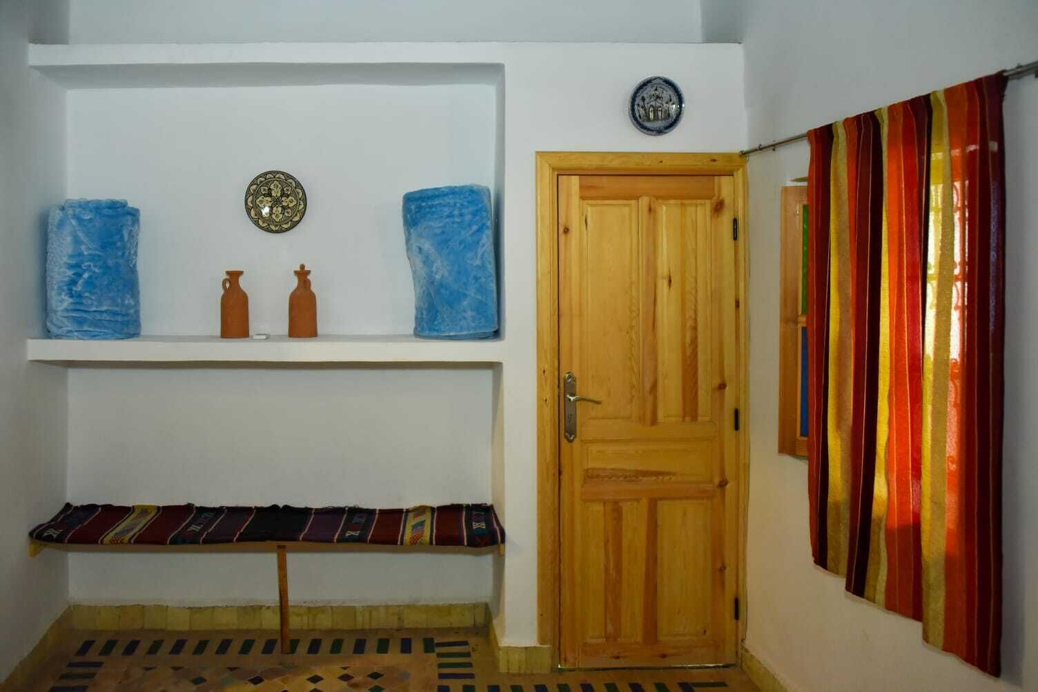 Una habitación con puerta de madera, una estantería con objetos decorativos y una cortina colorida.