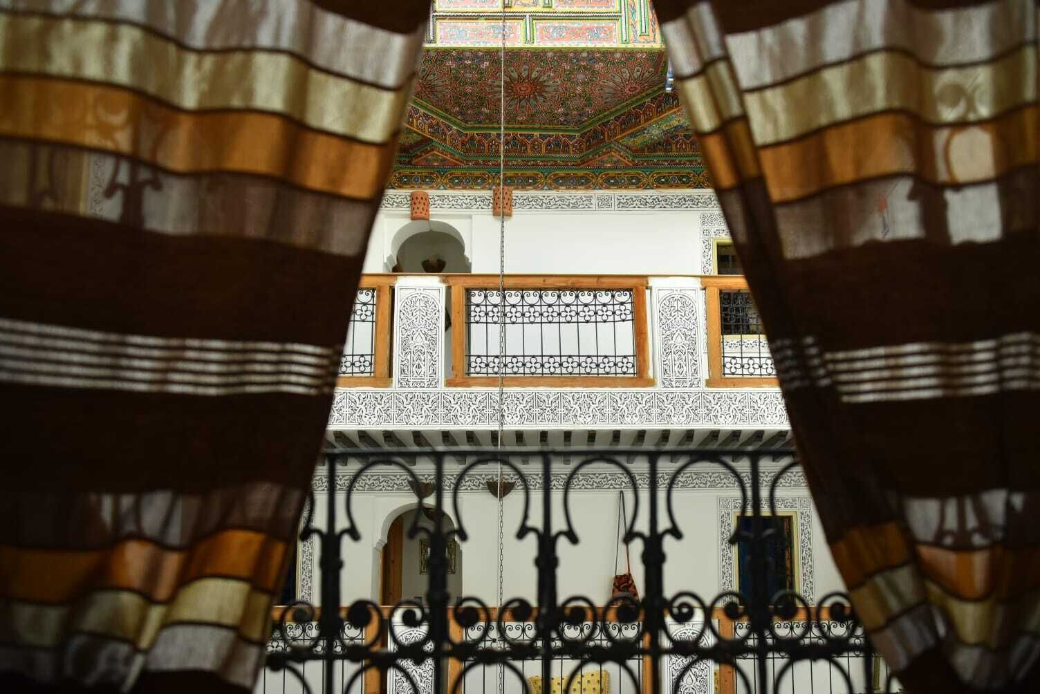 Visão através de cortina estampada, revelando uma varanda com grades ornamentadas e parede azulejada com desenhos intrincados.
