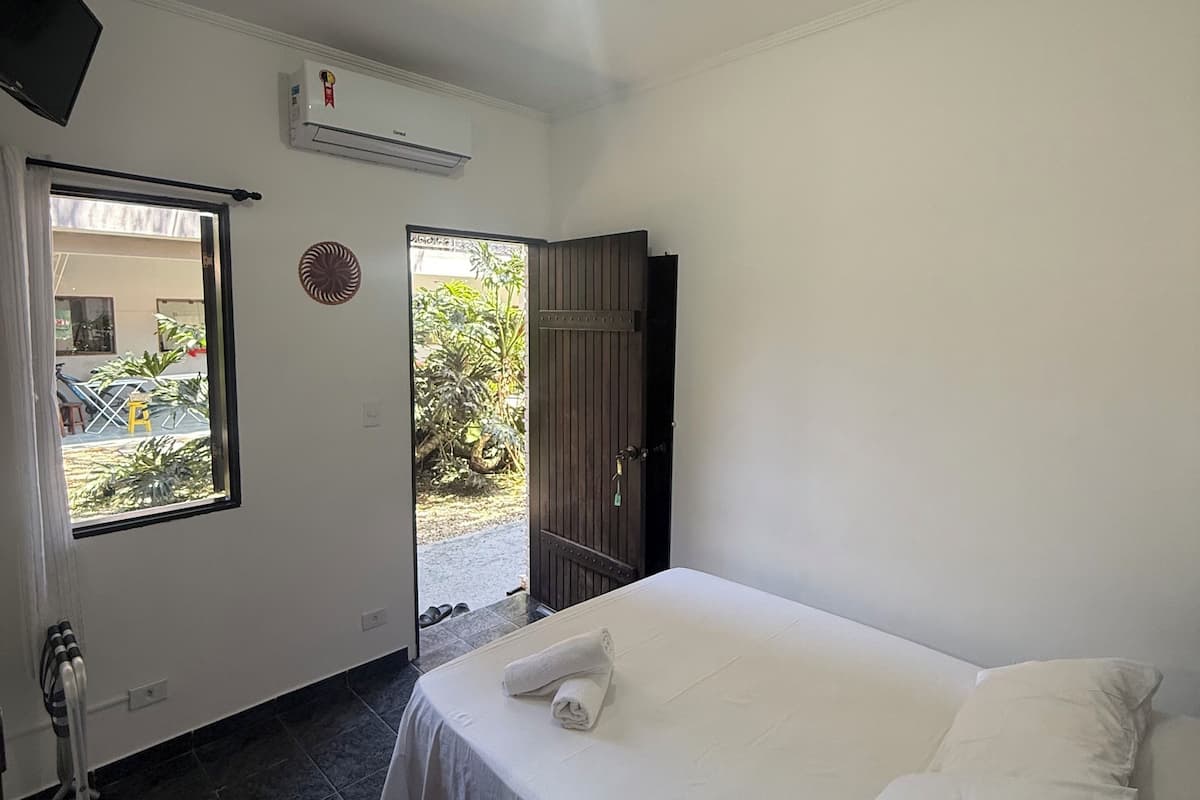 A hotel room with a bed, a window, an air conditioning unit, and a door leading to an outdoor area.