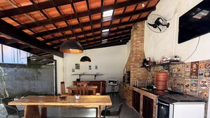 An outdoor dining area with a wooden table and benches, a brick oven, and a TV mounted on the wall. - Pousada Bahan (Ubatuba)