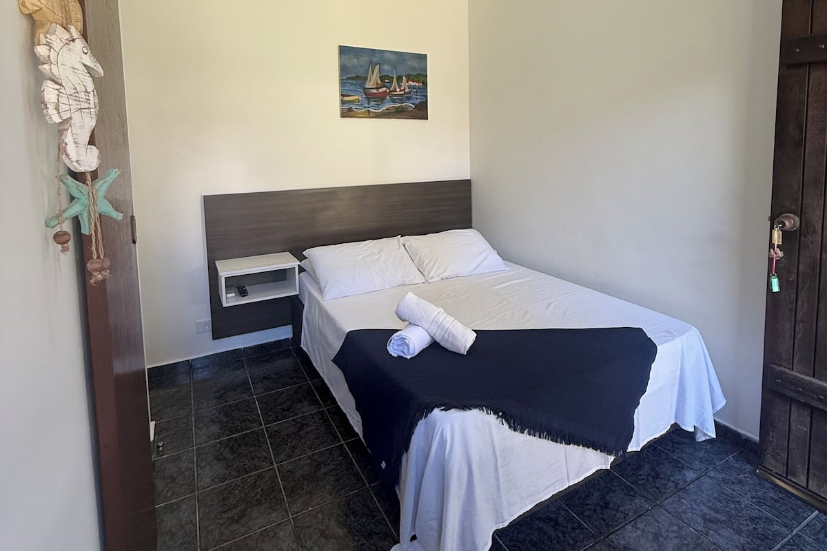 A neatly made bed with a white and black bedspread, a painting of a boat on the wall, and a wooden door on the right.