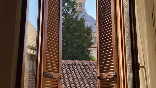 A view through an open window with wooden shutters, revealing a tiled roof and a building with a dome and statue in the background. - A view on Brescia's rooftops (Brescia)
