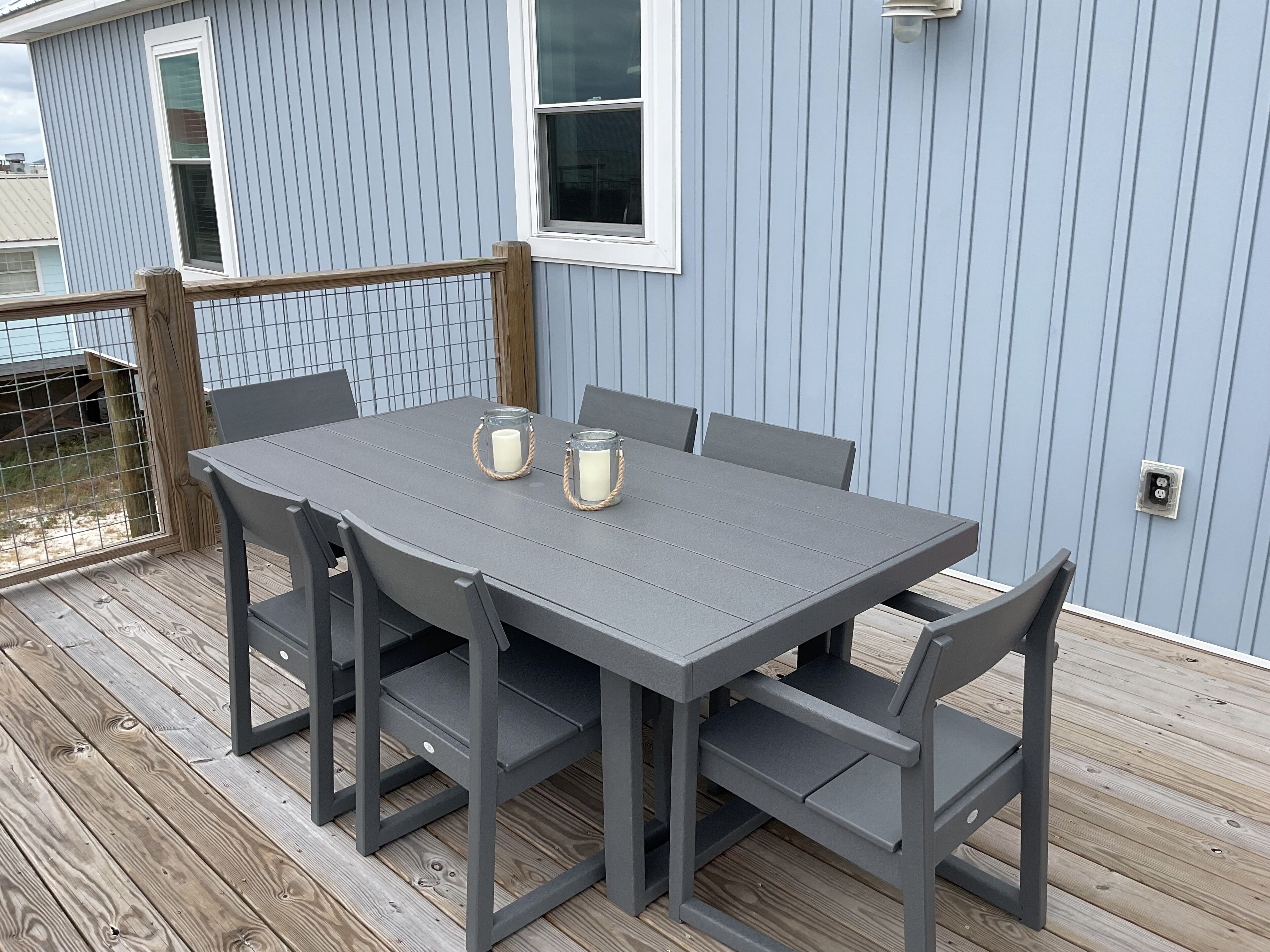 A gray metal table with six chairs on a wooden deck, set against a light blue building with vertical siding.