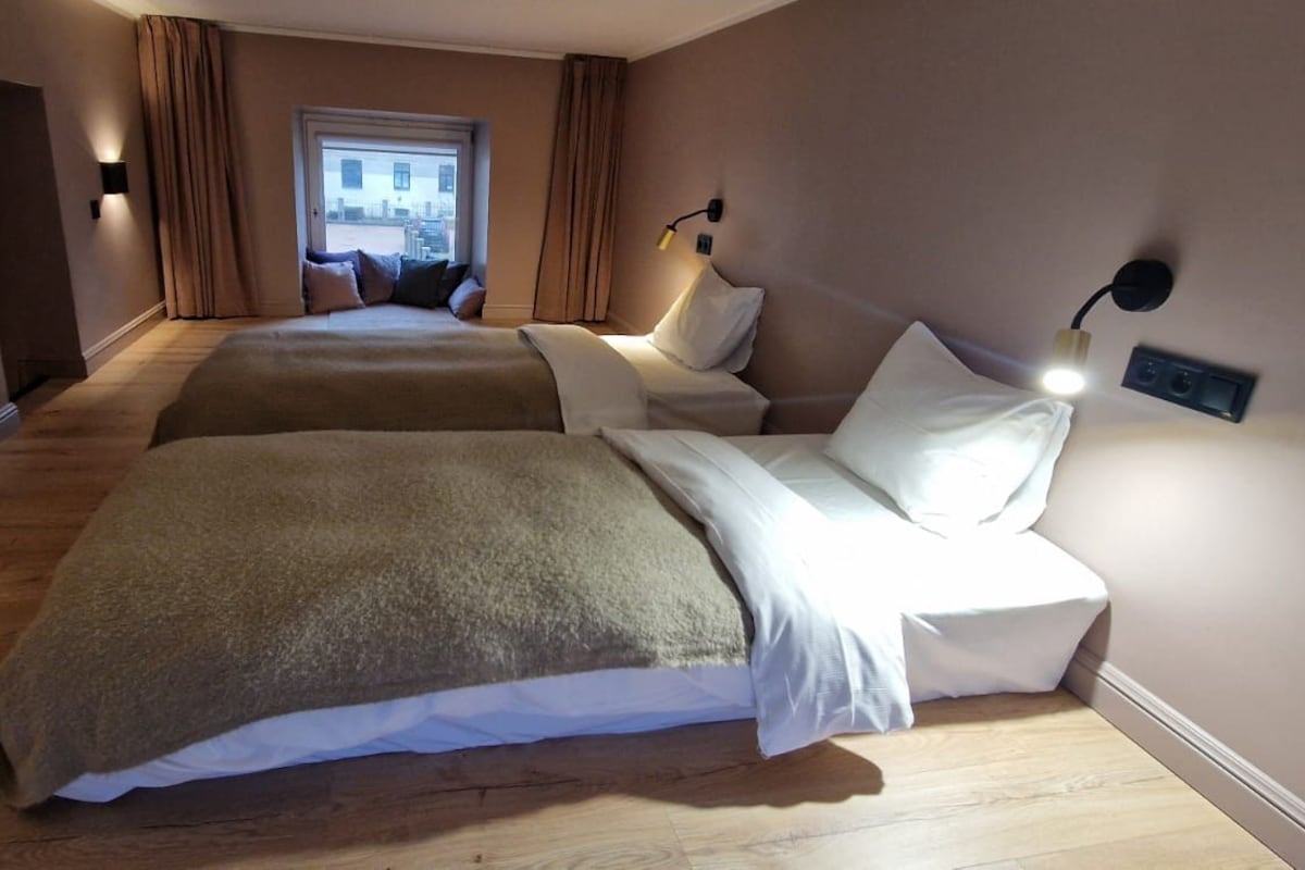 A hotel room with two beds, a large window with curtains, and a wall-mounted lamp.