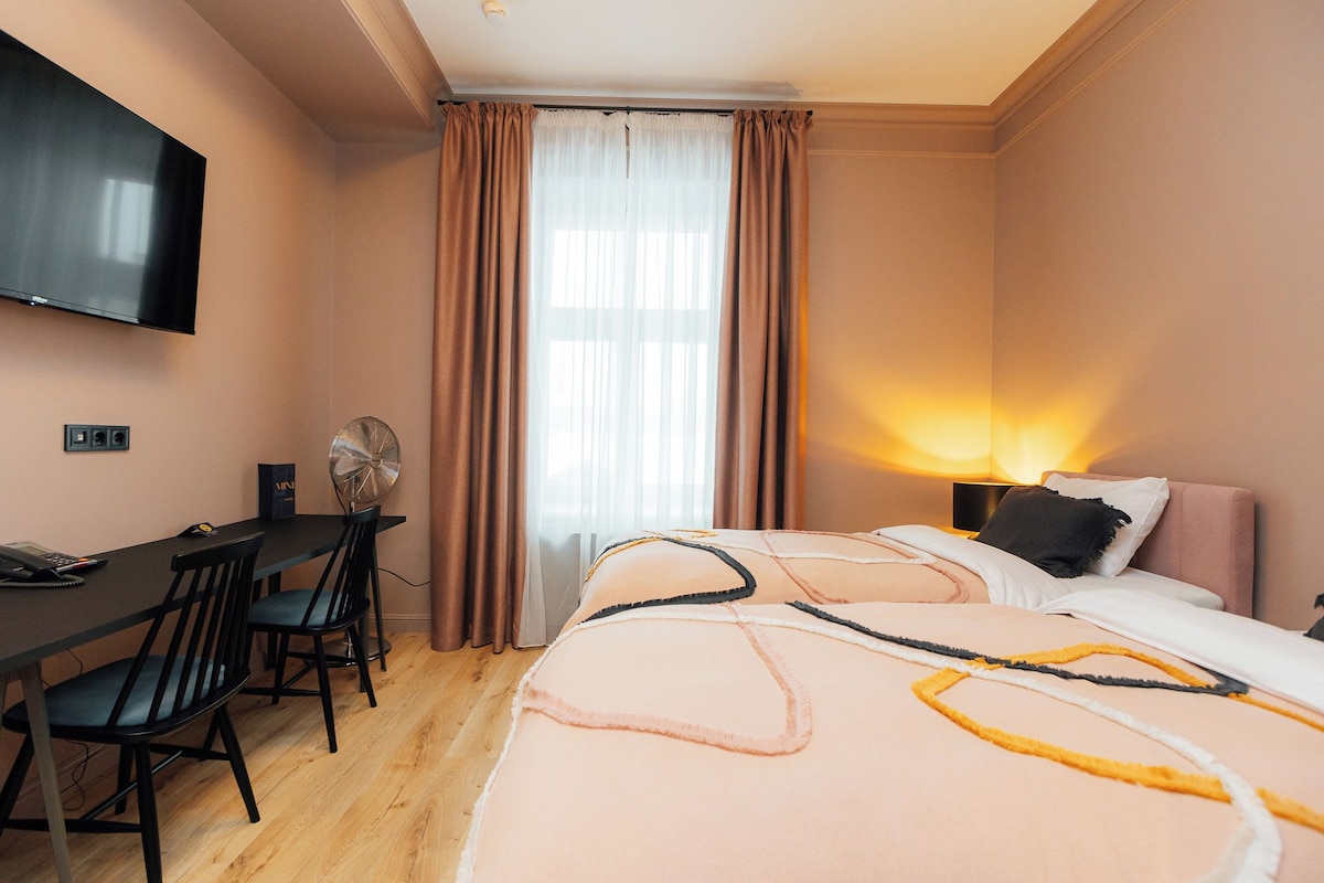 A hotel room with a bed, bedside table, lamp, TV, and a desk with chairs.