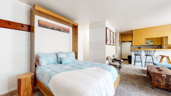 A bedroom with a bed, a wooden headboard, a nightstand, a desk, and a chair. There is a kitchenette with a refrigerator, microwave, and stove in the background.