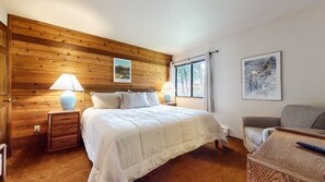Un dormitorio con cabecera de madera, cama con ropa de cama blanca, dos lámparas de noche, una ventana con vista a árboles y un cuadro en la pared.