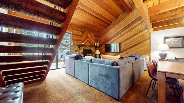 Interior acogedor de una cabaña con chimenea de piedra, un amplio sofá seccional gris y una mesa de comedor con sillas.