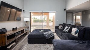 Un salon moderne doté d’un téléviseur à écran plat, d’un meuble de rangement en bois, d’un canapé noir agrémenté de coussins décoratifs et d’une grande porte-fenêtre coulissante donnant sur un espace extérieur.