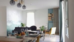 A modern living room with a dining area, featuring a sofa, a coffee table, and a dining table with chairs. There are hanging lights above the dining table and a large window providing natural light.