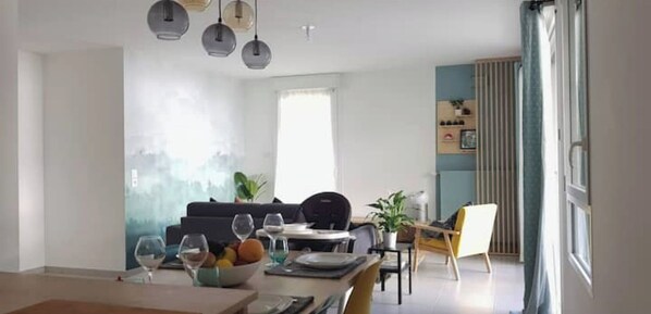 Un salon moderne avec un coin repas, comprenant un canapé, une table basse et une table à manger avec des chaises. Des luminaires suspendus sont placés au-dessus de la table à manger, et une grande fenêtre apporte une lumière naturelle.