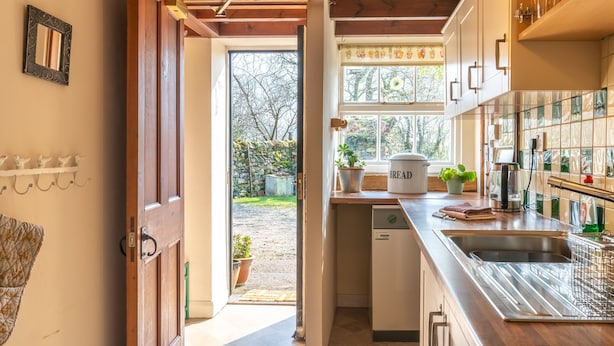 A kitchen with wooden cabinets, a built-in dishwasher, and a window with a view of the garden. - High Wallabarrow Farm Cottage -  Duddon Valley (Ulpha)