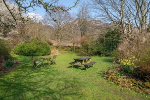 Ein Picknickbereich mit Tisch und Bänken in einem grünen Garten, umgeben von Bäumen und Sträuchern.