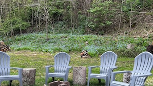 Tre Adirondack-stolar placerade runt en liten eldstad i ett skogsområde, med en hög med ved i närheten.