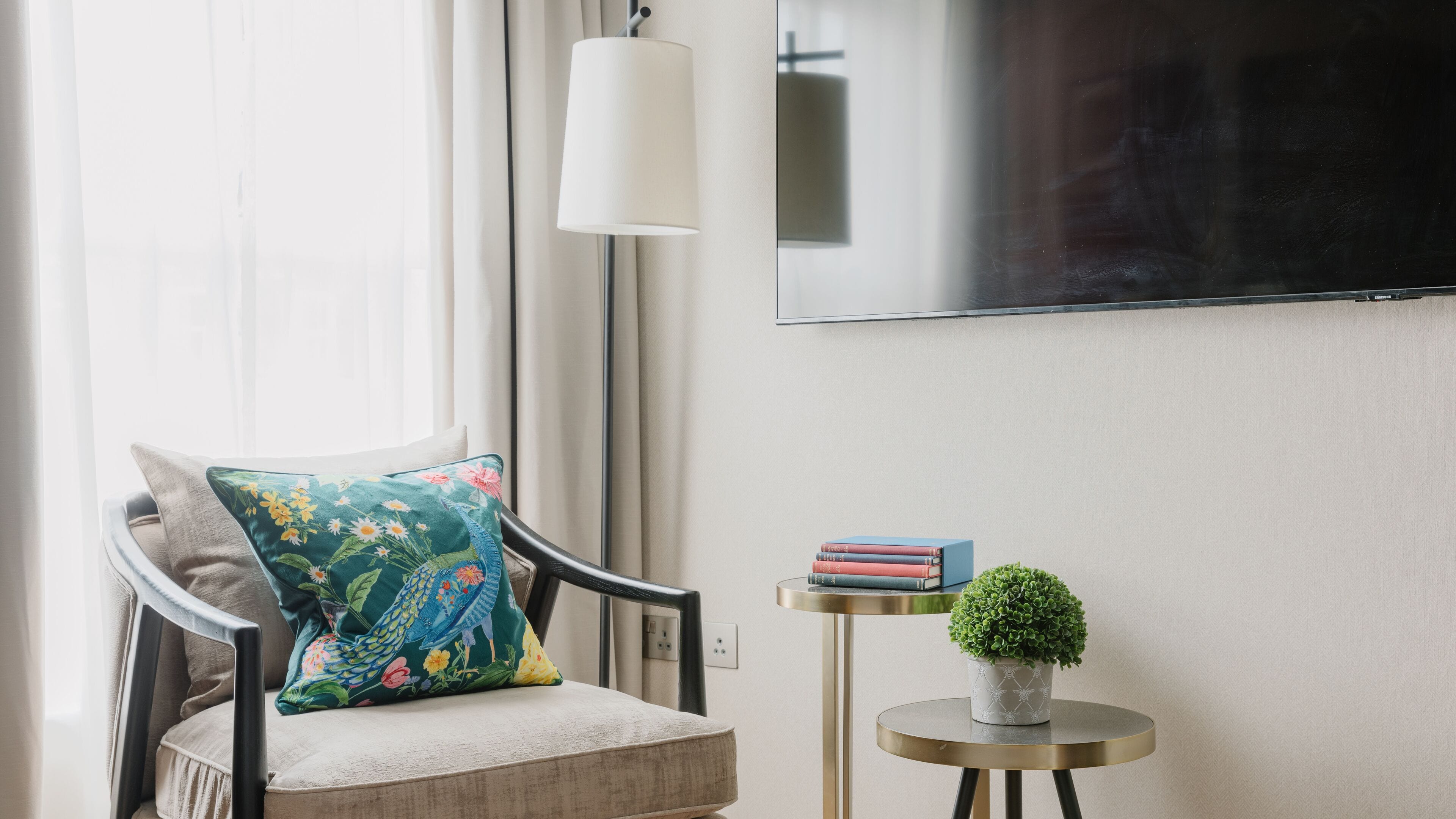 Uma sala de estar moderna com uma poltrona confortável, uma mesinha de apoio com livros e uma planta, e uma TV de tela plana na parede.