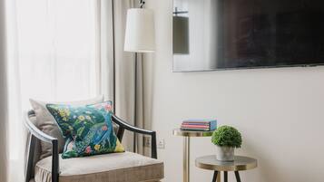 Uma sala de estar moderna com uma poltrona confortável, uma mesinha de apoio com livros e uma planta, e uma TV de tela plana na parede.