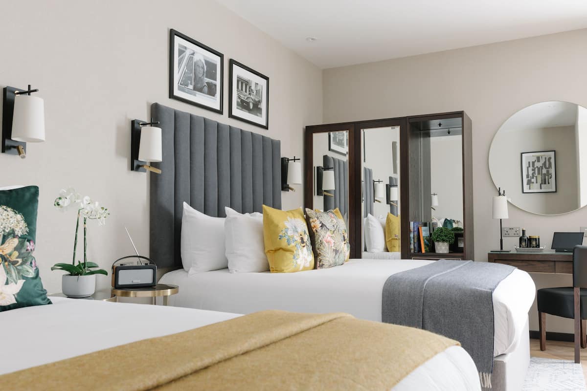 A modern hotel room with two beds, a desk, a chair, a round mirror, and a large window.