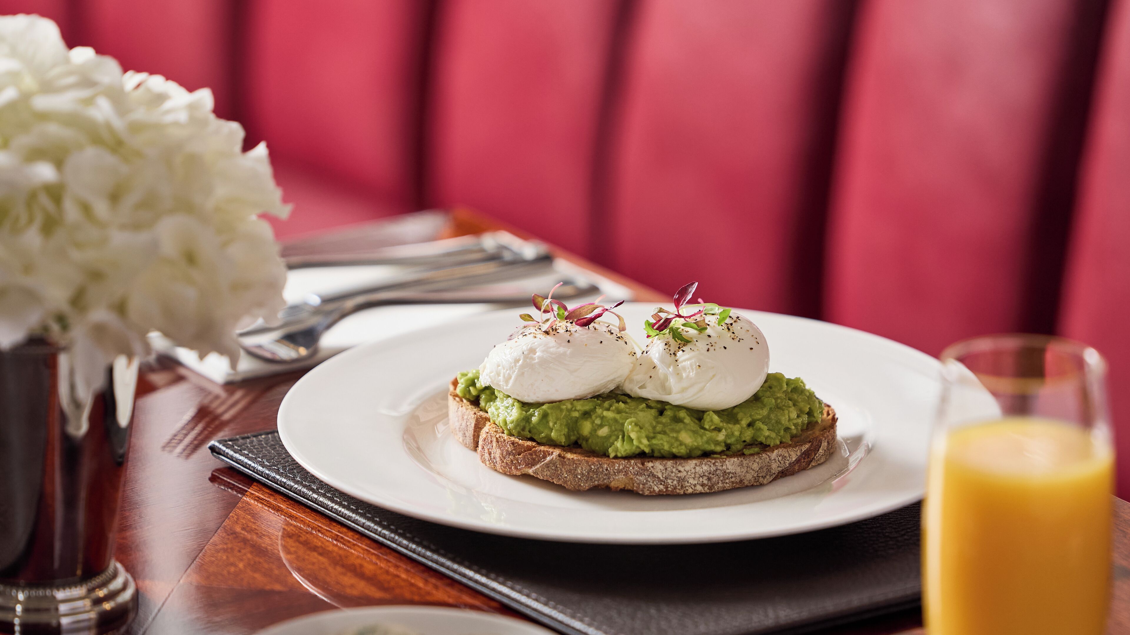 A breakfast plate with avocado toast and poached eggs, a glass of orange juice, and a vase of white flowers on a wooden table.