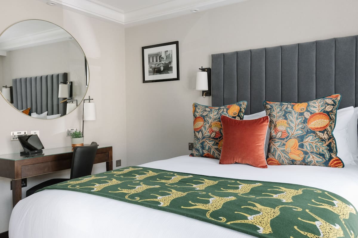 A hotel room with a large bed, a desk with a laptop, a mirror, a framed picture on the wall, and a potted plant.