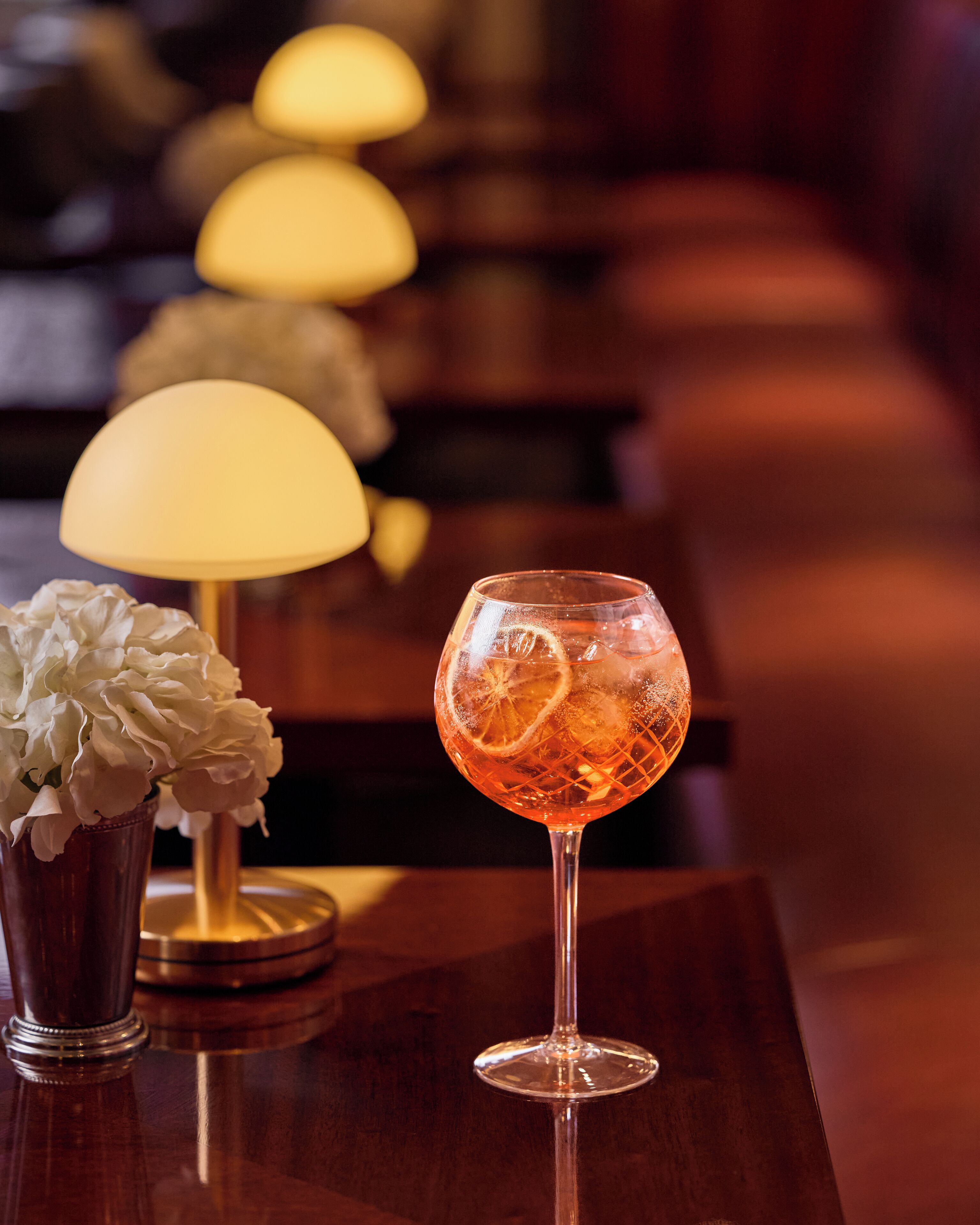 A glass of orange-colored drink with ice and a slice of orange, a white flower arrangement, and a lamp on a wooden table in a dimly lit room.