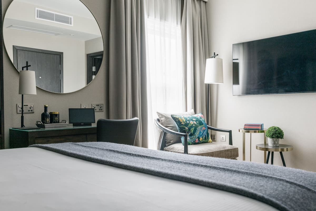 A modern hotel room with a large bed, a desk with a computer, a chair, a round mirror, a flat-screen TV, and a small side table with a plant.