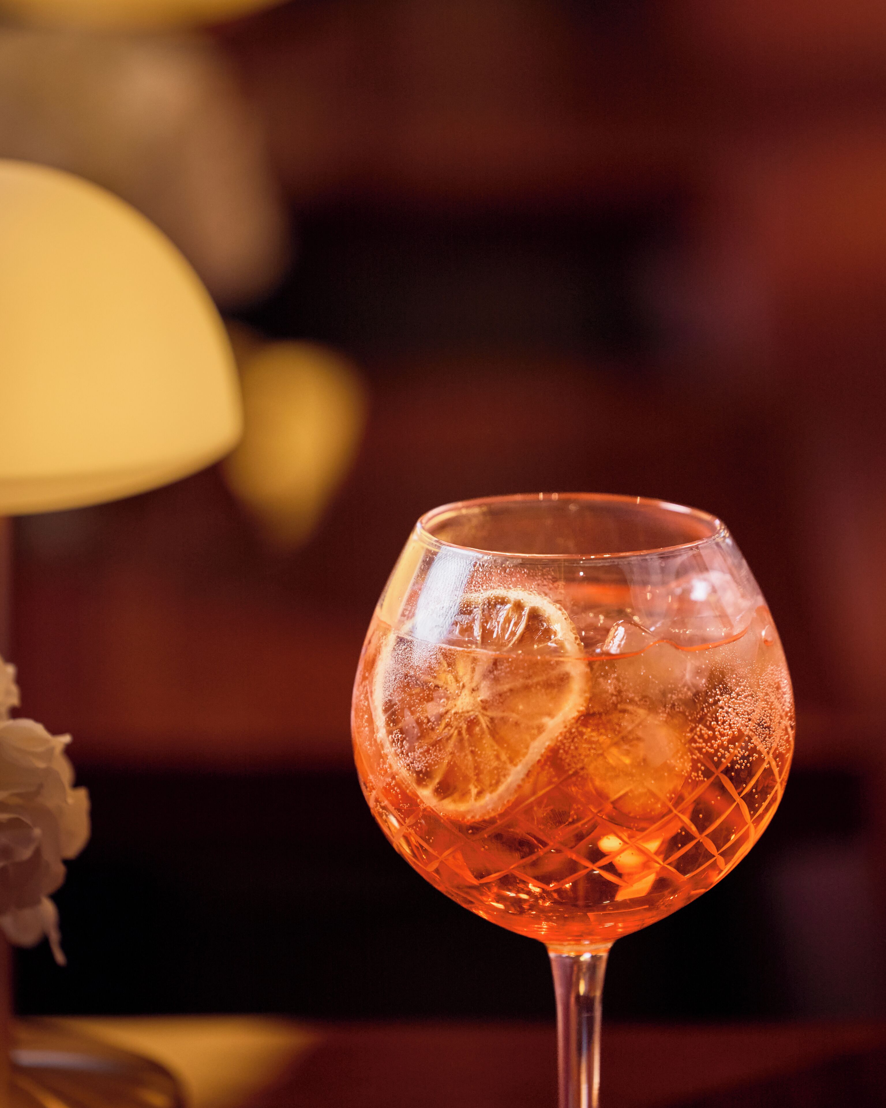 A glass of amber-colored beverage with a slice of citrus and ice cubes, set against a blurred background with warm lighting.