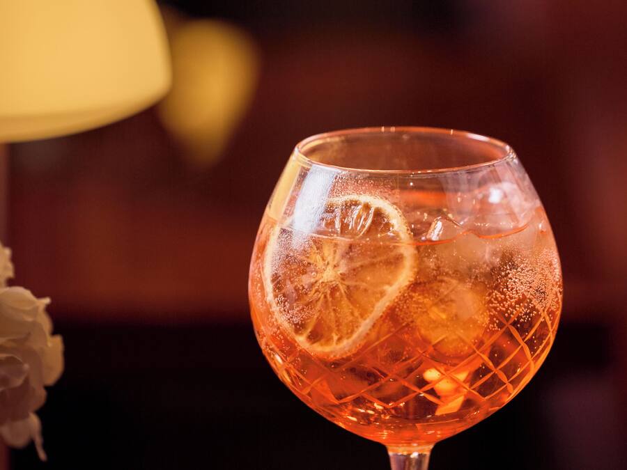 A glass of amber-colored beverage with a slice of citrus and ice cubes, set against a blurred background with warm lighting.