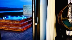 Une pièce dotée d’une porte-fenêtre coulissante donnant sur une terrasse en bois, où se trouvent un jacuzzi, un fauteuil suspendu et d’où l’on peut admirer l’océan au crépuscule.