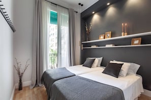 A modern bedroom with two beds, a large window with curtains, a dark accent wall, and a shelf with decorative items. - Bright Design Flat near Sagrada Familia (Barcelona)