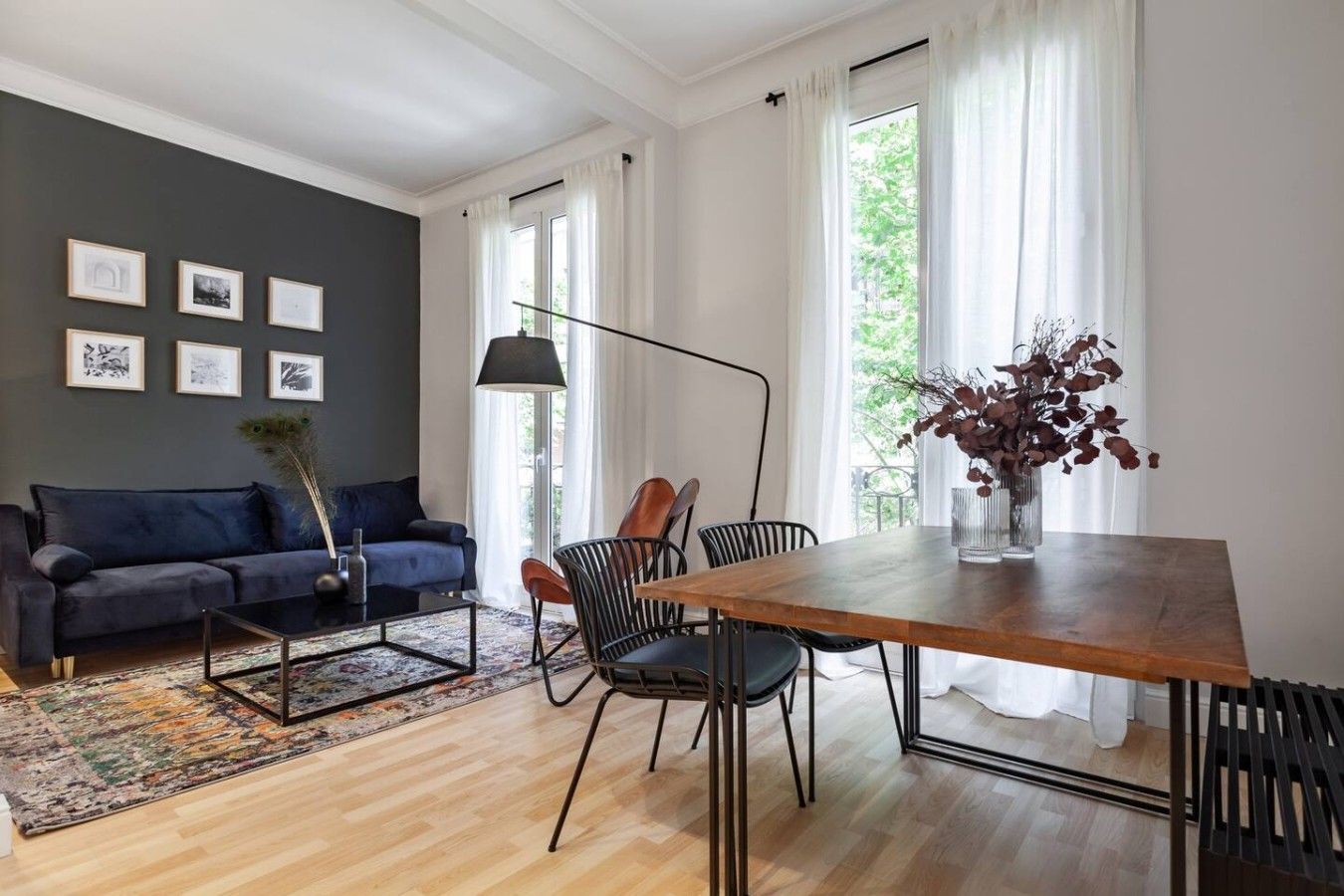 A modern living room with a dark blue sofa, a wooden dining table with chairs, a black floor lamp, and a large window with white curtains.