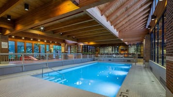 Una piscina interior con suelo de baldosas, vigas de madera en el techo y vistas al exterior a través de amplios ventanales.